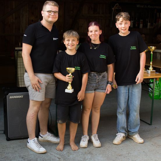 Fünf Personen stehen in einer Garage und lächeln für ein Foto. Sie tragen passende schwarze T-Shirts mit Text. Der Junge in der Mitte hält einen Pokal. Hinter ihnen sind Pokale und ein Tisch mit einem Lautsprecher zu sehen.