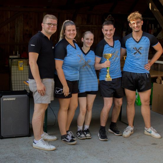 Fünf Personen stehen in einer Scheune zusammen, drei tragen blaue und schwarze Trikots, und zwei sind in schwarzen Hemden. Sie alle lächeln und posieren für ein Foto, wobei einer der Männer einen Pokal hält.