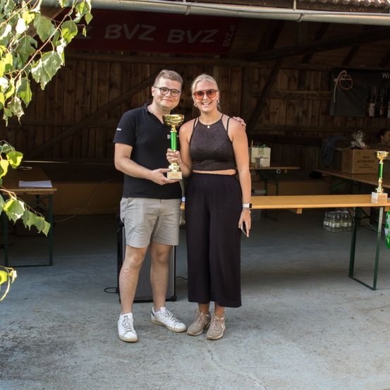 Ein Mann und eine Frau halten einen Pokal und lächeln in einem Holzschuppen mit einem Tisch, Flaschen und einem Banner, das BVz BVZ liest.