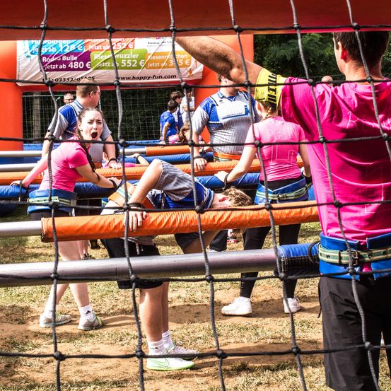 Eine Gruppe von Menschen spielt ein Spiel auf einem Feld mit einem Netz, zwei in pinkfarbenen Shirts lehnen sich über einen am Boden liegenden und ein anderer hält sich am Netz fest.