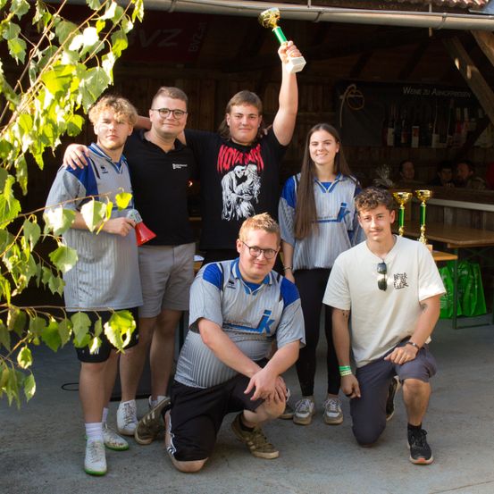 Eine Gruppe junger Erwachsener, in Sporttrikots gekleidet, posiert für ein Foto draußen. Einer hält eine Trophäe, und sie sind von Grünpflanzen und einem Gebäude mit Flaschen im Hintergrund umgeben.