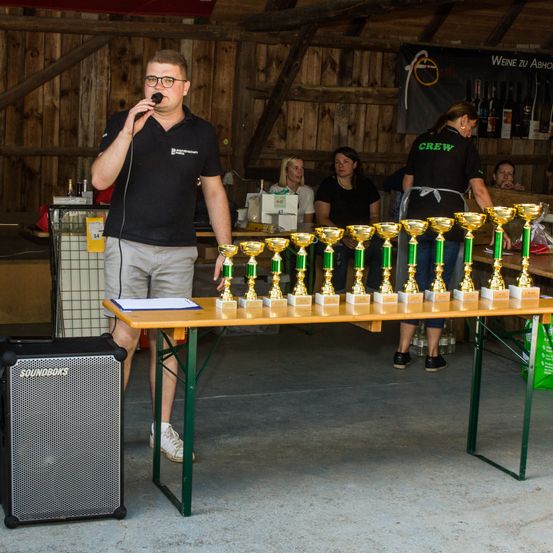 Ein Mann spricht in ein Mikrofon bei einer Veranstaltung. Vor ihm steht ein langer Tisch mit mehreren Trophäen. Dahinter stehen zwei Frauen in der Nähe eines Banners.