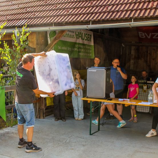 Ein Mann trägt einen großen Plastiksack vor einem Holzschuppen mit einem Schild, auf dem 'Cuhhaus Hauser' steht. Ein Mann mit Mikrofon spricht zu einer Gruppe von Menschen.