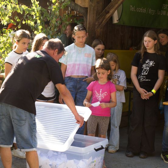 Ein Mann beugt sich über eine Plastikbox und füllt sie mit Gegenständen, umgeben von mehreren Mädchen, die ihn beobachten. Die Mädchen tragen verschiedene farbige Shirts und einige tragen Armbänder.