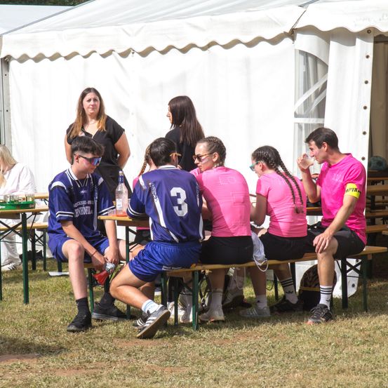 Eine Gruppe von Menschen, darunter zwei Frauen und vier Männer, sitzen auf Bänken unter einem weißen Zelt. Zwei der Männer tragen blaue Trikots, während die anderen beiden in Pink sind. Ein Mann in einem pinken Hemd trinkt aus einem Becher.