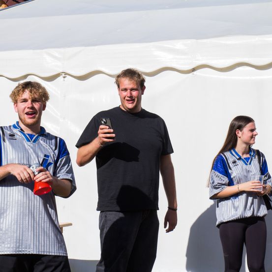 Drei Personen stehen in einem Zelt, ein Mann auf der linken Seite hält eine Flasche, der Mann in der Mitte hält ein Telefon und die Frau auf der rechten Seite hält einen Becher.