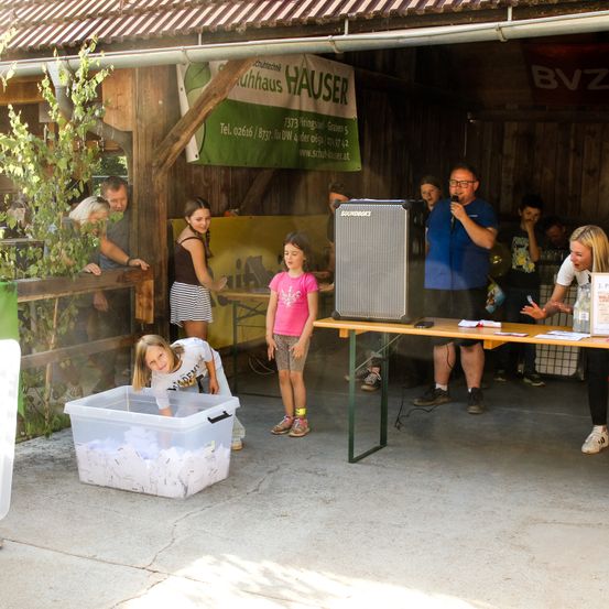 Eine Gruppe von Menschen versammelt sich um einen Tisch mit einem Lautsprecher, ein Mädchen schaut in eine Lagerbox und ein Mann spricht in ein Mikrofon.