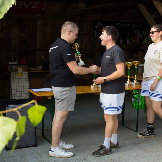 Zwei Männer, einer mit einer Brille und der andere mit einem grünen Armband, schütteln sich die Hände. Sie stehen vor einem Tisch mit Pokalen. Hinter ihnen steht eine Frau mit Sonnenbrille.