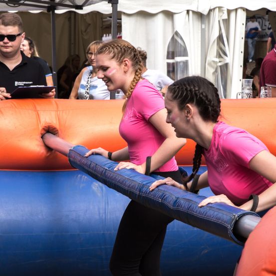 Zwei Frauen in rosa Shirts nehmen an einer Veranstaltung teil, eine schiebt einen blauen Schlauch, die andere beobachtet, mit Zuschauern im Hintergrund.