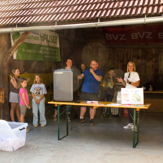 Ein Mann steht in einer Scheune an einem Tisch und spricht in ein Mikrofon, während eine Frau neben ihm steht und der Menge zugewandt ist. Vor ihnen versammeln sich Kinder und andere Erwachsene. An der Wand hängt ein Banner.