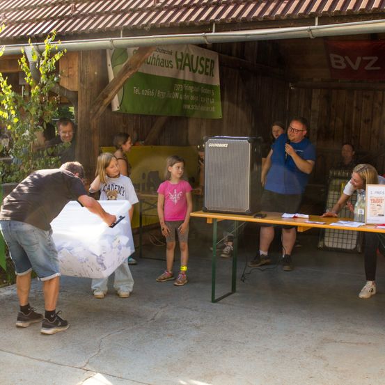 Eine Gruppe von Menschen in einer Scheune mit einer Bühneneinrichtung, eine Person hält eine Box, während eine andere in ein Mikrofon spricht. Ein Banner hängt im Hintergrund.