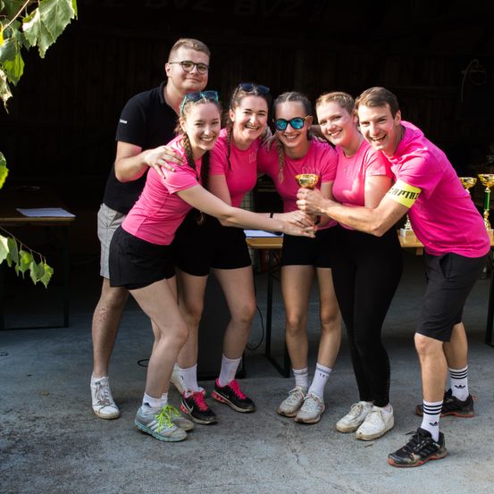 Eine Gruppe lächelnder junger Menschen in rosa Shirts und schwarzen Shorts steht in einer Reihe. Sie halten eine Trophäe in der Mitte. Ein Mann steht hinter ihnen. Sie sind wahrscheinlich Athleten. Es gibt Tische und Pflanzen hinter ihnen.