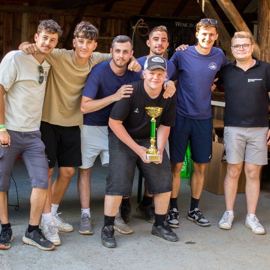 Sechs Männer stehen in einer Reihe, lächeln und posieren für ein Foto. Der Mann in der Mitte hält einen goldenen Pokal. Der Hintergrund zeigt eine Holzstruktur.