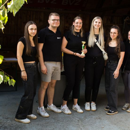 Sechs Personen in Schwarz stehen in einer Gruppe. Eine Frau in der Mitte hält eine Trophäe. Sie stehen vor einem Holzgebäude.