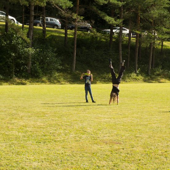 Zwei Kinder auf einem Rasenfeld, eines macht einen Handstand und das andere hält einen grünen Ring. Dahinter sind mehrere Autos auf einem Hang geparkt.