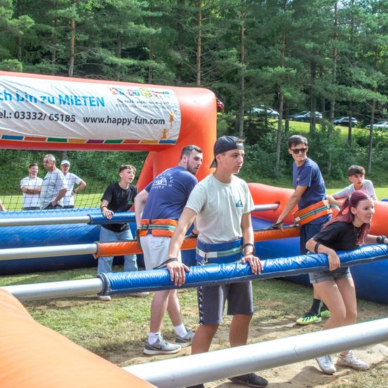 Eine Gruppe von Menschen spielt in einem aufblasbaren Hindernisparcours. Sie tragen Gurte und scheinen einen blauen Stab zu schieben. Bäume und geparkte Autos sind im Hintergrund.