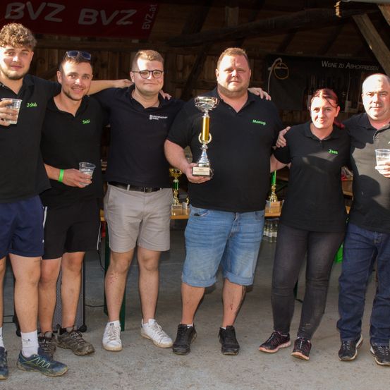 Fünf Erwachsene stehen vor einem Banner und posieren für ein Foto mit Gläsern in den Händen. Der Mann in der Mitte hält einen Pokal.