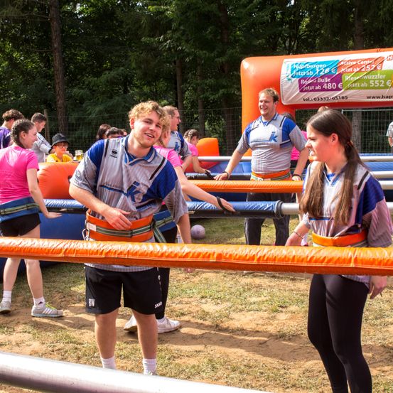 Eine Gruppe von Menschen in Trikots nimmt an einem Outdoor-Spiel mit einem aufblasbaren Hindernis teil. Eine Frau und ein Mann stehen vor einer orangefarbenen Röhre und lächeln. Dahinter hält ein Mann einen Ball und eine Frau lacht. Bäume und ein Zaun sind im Hintergrund.