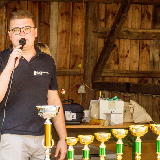 Ein Mann mit Brille spricht in ein Mikrofon in einer Scheune, mit Pokalen auf dem Tisch vor ihm. Dahinter befinden sich Holzwände und ein Tisch mit Kisten.