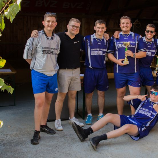 Eine Gruppe junger Männer in Sportuniformen, möglicherweise ein Team, posiert für ein Foto mit einer Trophäe. Sie stehen in einer Reihe, einige mit Armen auf den Schultern der anderen.