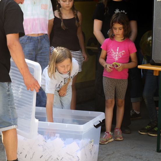Ein Mann hält einen durchsichtigen Plastikbehälter voller Papiere, während ein Mädchen hineinschaut. Hinter ihnen schauen Kinder auf den Behälter. Ein Mädchen trägt ein rosa T-Shirt.