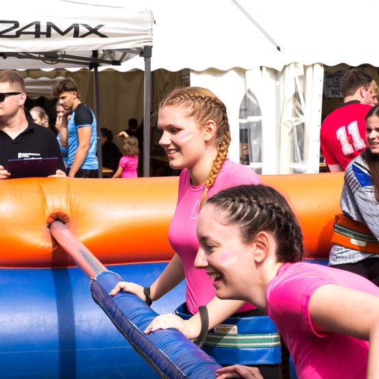 Zwei junge Frauen, die rosa T-Shirts tragen, lachen und halten sich an einem blauen Rohr in einem orangefarbenen aufblasbaren Hindernisparcours fest. Ein Mann in einem schwarzen Hemd steht und beobachtet.