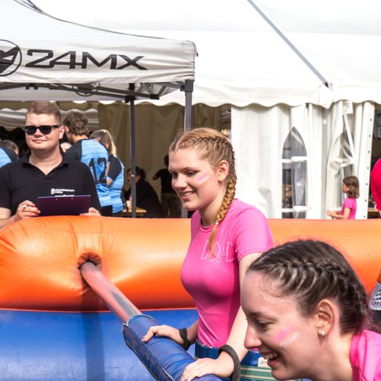 Zwei Frauen lächeln in einem aufblasbaren Hüpfhaus. Ein Mann mit einer Brille und einem Tablet steht hinter ihnen, unter einem Zelt mit dem Z4MX-Logo.