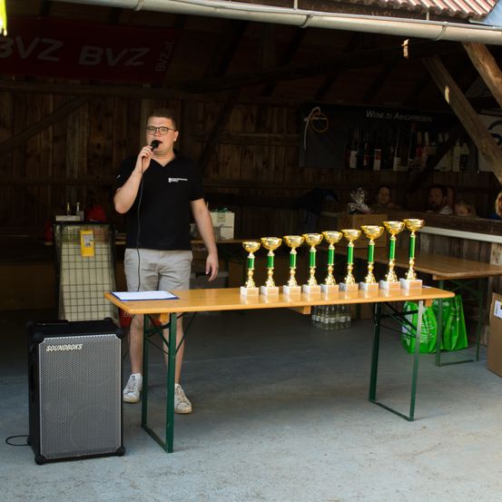 Ein Mann spricht in ein Mikrofon, während er vor einem Tisch mit mehreren Trophäen steht. Er befindet sich in einem Raum mit Holzwänden.