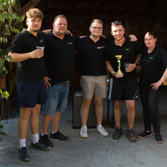 Fünf Personen, die gleiche schwarze Hemden tragen, stehen zusammen und halten einen Pokal. Sie sind draußen, mit einem Banner, das BVz hinter ihnen liest.