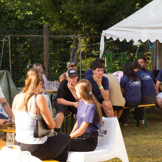 Eine Gruppe von Menschen sitzt zusammen unter einem Zelt, einige tragen Freizeitkleidung und andere tragen passende blaue Shirts. Sie unterhalten sich miteinander, während sie Getränke in den Händen halten.