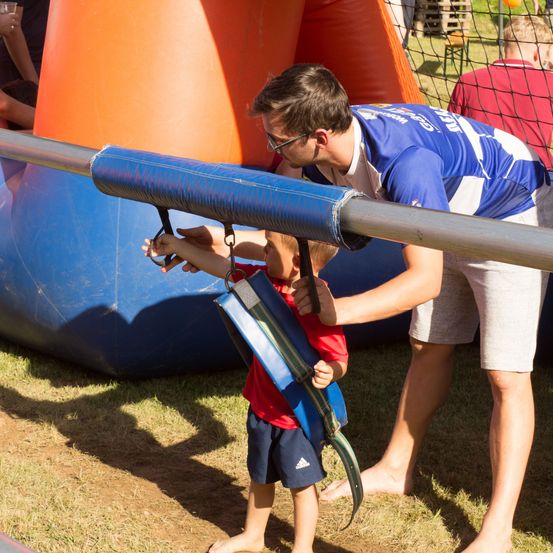 Ein Mann hilft einem kleinen Jungen, der einen Gurt trägt, der an einem blauen Objekt befestigt ist, möglicherweise eine Attraktion bei einer Outdoor-Veranstaltung.