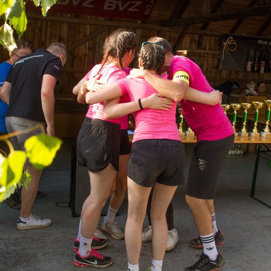Eine Gruppe von Menschen in rosa Shirts und schwarzen Shorts steht zusammen vor einem Tisch mit Trophäen. Zwei Männer umarmen die Frauen von hinten. Ein anderer Mann in einem schwarzen Shirt beobachtet sie.