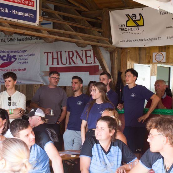 Eine Gruppe von Menschen in blauen T-Shirts steht in einem Raum, vor einem Banner mit der Aufschrift 'THURNER'. Einige tragen Brillen und halten Tassen, während eine Treppe und eine Holzwand im Hintergrund zu sehen sind.