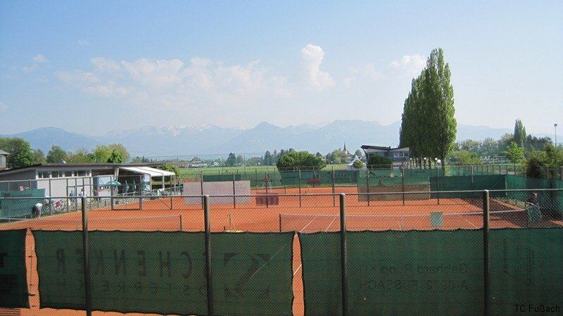Freiluft-Tennisplätze mit Blick auf die Berge, eingezäunt durch einen grünen Zaun. Ein Gebäude mit Vordach und Menschen in der Nähe.