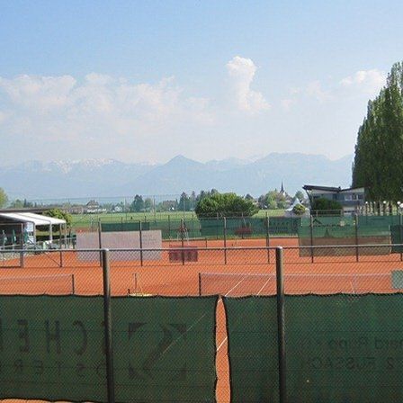 Freiluft-Tennisplätze mit Blick auf die Berge, eingezäunt durch einen grünen Zaun. Ein Gebäude mit Vordach und Menschen in der Nähe.