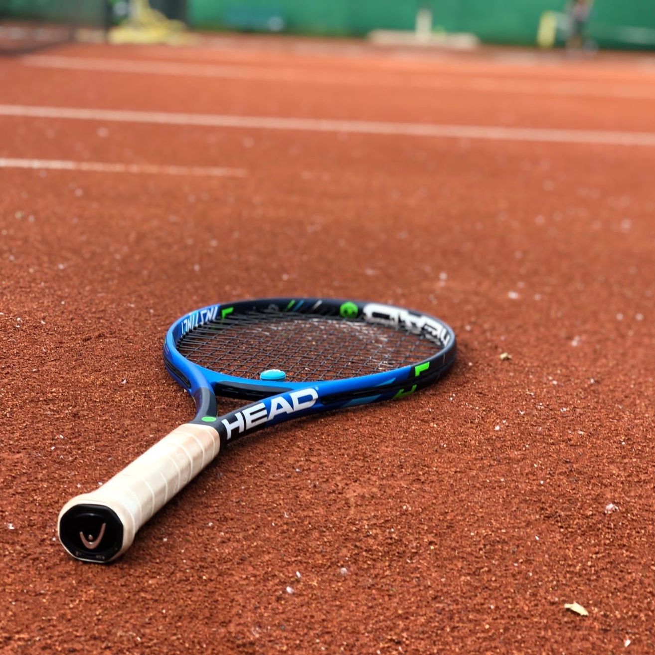 Ein blauer und weißer HEAD-Tennisschläger liegt auf dem Sandplatz mit einem grünen Ball auf dem Schläger. Der Boden ist mit Sandstaub bedeckt.