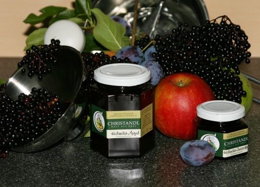 Auf einem Tisch sind verschiedene Früchte und Marmeladen zu sehen. Gläser mit schwarzer Holunder- und Pflaumenmarmelade sind vorhanden, zusammen mit frischen schwarzen Holunderbeeren, Pflaumen und Äpfeln. Ein Edelstahl-Messbecher und eine Schüssel mit Eiern sind ebenfalls auf dem Tisch.