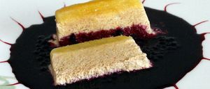 Zwei Stücke Käsekuchen mit einer Schicht Beerenkompott liegen auf einem weißen Teller mit schwarzer Beerensauce. Das Dessert ist mit roten Linien verziert.