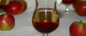 Ein klares Glas mit goldener Flüssigkeit steht auf einem weißen Tisch mit Äpfeln im Hintergrund.
