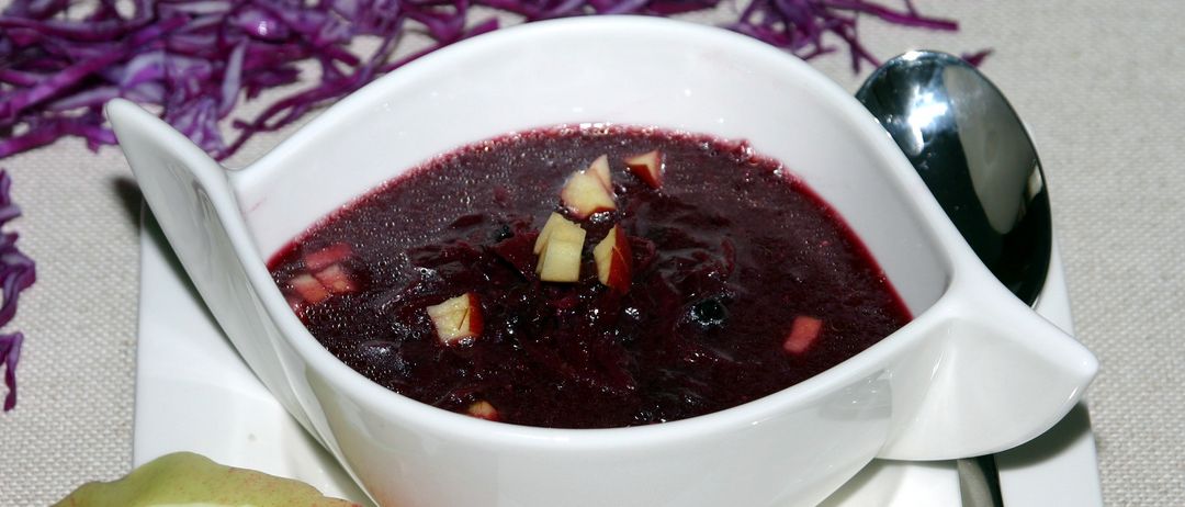 Ein weißer Teller mit einer dunkelroten Suppe mit Apfelstücken, dazu ein Löffel und lila Kraut auf dem Tisch.