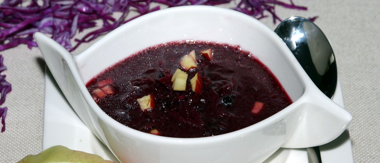Ein weißer Teller mit einer dunkelroten Suppe mit Apfelstücken, dazu ein Löffel und lila Kraut auf dem Tisch.