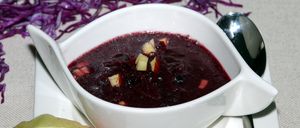 Ein weißer Teller mit einer dunkelroten Suppe mit Apfelstücken, dazu ein Löffel und lila Kraut auf dem Tisch.