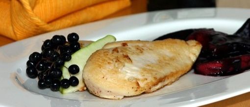 Ein weißer Teller mit einem Stück Hähnchen, geschnittenen Gurken und schwarzen Johannisbeeren, mit einem Glas Marmelade im Hintergrund.