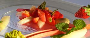 Ein Teller mit einer lebhaften Mischung aus geschnittenen Erdbeeren, Rhabarber und Minzblättern, überzogen mit einer roten Sauce. Das Gericht ist mit einer Rhabarberscheibe und einem Minzblatt garniert.