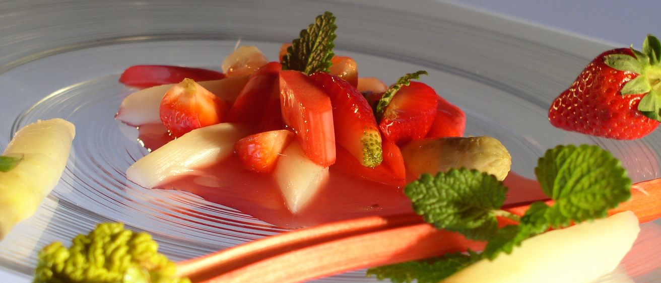 Ein Teller mit einer lebhaften Mischung aus geschnittenen Erdbeeren, Rhabarber und Minzblättern, überzogen mit einer roten Sauce. Das Gericht ist mit einer Rhabarberscheibe und einem Minzblatt garniert.