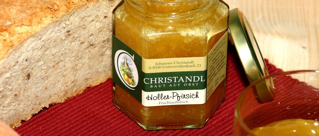 Ein Glas Christandl Holler-Pfirsich-Marmelade ist auf einem roten Tuch geöffnet, mit einer Scheibe Brot und einem Glas in der Nähe.