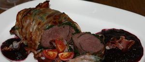 Ein Teller mit Wildschweinfleisch, das in Speck und Kräutern gewickelt ist, serviert mit Rotweinsauce und Kirschtomaten, begleitet von einer Schüssel Rotkohl.