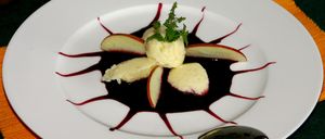 Ein weißer Teller mit einem Dessert bestehend aus Apfelscheiben, einer Sahnehaube und roter Sauce. Obenauf befindet sich ein Minzzweig.