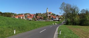 Eine malerische Straße, umgeben von üppigem Grün, führt zu einem malerischen Dorf mit rot gedeckten Häusern und einer Kirche mit Glockenturm.