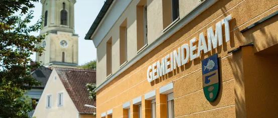Ein gelbes Gebäude mit dem Wort Gemeindeam in Weiß auf der Vorderseite. Ein Kirchturm ist im Hintergrund sichtbar.